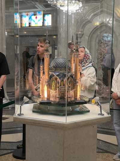 The image depicts the interior of a museum, likely in the city of Usady, Russia, showcasing a detailed architectural model of a mosque displayed within a glass case. A young man with short brown hair, wearing a grey t-shirt, stands to the left of the display, looking towards it. To the right of the model, a woman with a colorful headscarf and a white jacket gazes upwards at the mosque model. She is wearing a watch on her left wrist. Reflected in the glass are other museum visitors and architectural elements of the building. The mosque model itself is ornate, featuring multiple minarets, domes, and intricate golden detailing. The display case is lit by small spotlights. The surrounding environment appears to be a well-lit hall with marble finishes and decorative arches visible in the background.