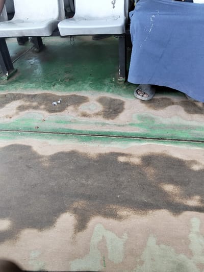 The scene depicts a ground-level view within an establishment in Mkomani ward, Kenya, likely a waiting area or public space. The floor is composed of a light green surface, heavily worn and discolored in patches, revealing a darker grey-brown substrate. Numerous irregular, dark, damp or stained areas are visible across the floor, along with scattered small debris and a crumpled white paper object. Two light grey plastic seats with black metal frames are positioned in the upper portion of the frame. To the right, the lower portion of a person is visible, clad in a dark blue garment that drapes over their legs, with one foot exposed, wearing a grey open-toed slide sandal. Beneath the seats on the left, the lower legs and dark footwear of another individual are partially discernible. No specific actions or interactions are depicted; the individuals appear to be seated or stationary.