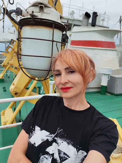 A woman with short, peachy-pink hair and bold red lipstick poses for a selfie on the deck of a ship. She is wearing a black t-shirt with a white graphic print. In the background, part of a large industrial light fixture with a metal cage and chain is visible, along with yellow machinery and the white and red superstructure of the ship. The deck is painted green. The lighting suggests it is daytime, and the weather appears overcast. The woman's expression is pleasant. The scene takes place in Kurdiug, Russia.