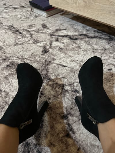 The image shows a close-up, top-down view of a person's feet wearing black heeled ankle boots. The boots are made of a suede-like material and have zippers on the inner sides. The person is standing on a patterned carpet with grey and white marbling. In the background, a wooden furniture piece, possibly a dresser or cabinet, is visible, along with a stack of books on the floor. There is no identifiable location in the city of Kalininskaya, Russia.