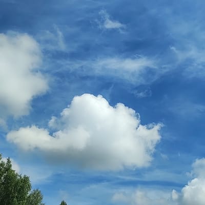 The image shows a blue sky with scattered white clouds. On the lower left, the tops of green trees are visible. The scene appears to be an outdoor environment, possibly a park or a natural area. There are no people, animals, or man-made structures clearly identifiable in the image, aside from the suggestion of trees, which could indicate the presence of a town or settlement. The lighting suggests it is daytime, with the sun likely overhead. The weather appears fair with a mix of cloud cover and clear sky. There is no specific indication of the location within Khokhlyata, Russia.