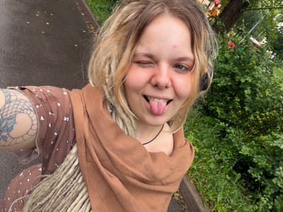 A young woman with blonde hair and dreadlocks is taking a selfie outdoors. She is wearing a brown floral dress and a light brown scarf. She is winking and sticking out her tongue, with a playful expression. Her left arm, which has a tattoo, is visible. In the background, there is a wet paved area and lush green foliage, suggesting a park or a residential area with trees. A faint hint of playground equipment can be seen in the distance. The scene appears to be in the Vostochny District of Russia.