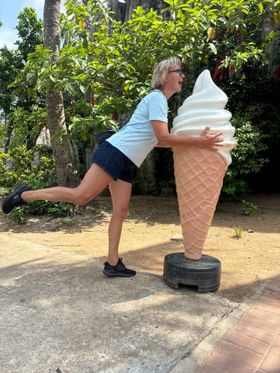 An adult Caucasian female with blonde hair and glasses leans against a very large ice cream cone statue. The statue is a light brown waffle cone topped with white soft-serve ice cream. The woman is wearing a light blue t-shirt and dark denim shorts. She is holding onto the ice cream cone with both hands and has her mouth open as if to take a bite. Her left leg is extended behind her. The scene is outdoors, with a sandy ground and a concrete pathway. Lush green trees with red ribbons tied to their branches are in the background, along with a rocky cliff face. The location is identified as Nanwan Village, China.
