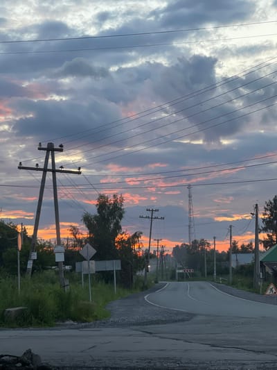 The image depicts a rural road in Druzhinino, Russia, during a sunset. The sky is filled with dramatic clouds displaying hues of pink, orange, and grey. Power lines crisscross the frame, supported by wooden utility poles. Trees line the sides of the road, some bare and others with foliage. A paved road curves into the distance, with a smaller gravel road branching off to the left. Road signs are visible, including a directional sign indicating a curve and a warning sign depicting a construction worker. There are no people visible in the image. The overall scene suggests a quiet, end-of-day atmosphere in a village setting.
