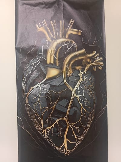 The media file is a photograph of a large, black banner featuring an artistic depiction of a human heart. The heart is rendered in a stylized manner, with golden, vein-like structures intricately branching across its surface. The background of the banner is dark and textured, creating a dramatic contrast with the luminous golden details of the heart. The image is abstract and somewhat surreal, evoking a sense of both organic form and intricate design. There is no discernible text or any indication of a specific time of day, weather, or emotions. The location context is Sagarejo, Georgia, but this information is external to the image itself and not visible within it.