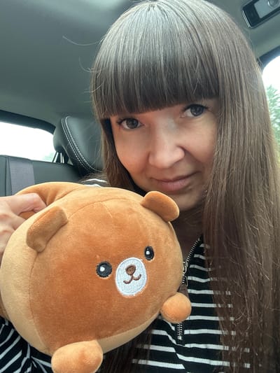 A young woman with long brown hair and bangs is holding a round, plush teddy bear in the front passenger seat of a car. She is looking directly at the camera with a slight smile. The interior of the car is visible, including a black leather seat with white stitching and the car's roof. The background outside the car shows greenery, suggesting a location outside of a city, potentially a rural area or a park within a town. The location is identified as Novaya Kazmaska, Russia.