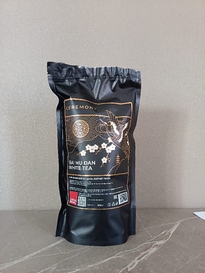 A black, stand-up pouch of "CEREMONY BAI MU DAN WHITE TEA" is prominently displayed, likely in an indoor setting. The pouch features an intricate gold and white design against its matte black surface. The label illustrates a stylized sun or moon with an ornate pattern, a majestic crane bird in flight, delicate white cherry blossoms on a branch, and a small pagoda structure on a mountainous backdrop.

Text visible on the packaging includes "CEREMONY," "BAI MU DAN," and "WHITE TEA" in English. Below this, in Cyrillic, it reads "ЧАЙ БЕЛЫЙ БАЙ МУ ДАНЬ (БЕЛЫЙ ПИОН)," which translates to "White Bai Mu Dan Tea (White Peony)." Further down, the weight "100 г" (100 grams) is clearly marked. A red square logo partially visible on the bottom left appears to be "ORIENTAL BAZAR." There are also several small lines of text, QR codes, and the "EAC" conformity mark, indicating product information and certifications.

The pouch is resting on a marbled, light grey and white countertop or table surface, contrasting with a plain, light grey wall in the background. The lighting is even, suggesting an indoor, controlled environment, possibly a product photography setup. No human figures or activities are present. The scene is a simple product shot, showcasing the tea packaging from Tsibanobalka, Russia.