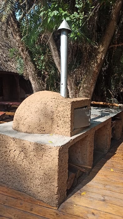 An outdoor pizza oven, made of earthen material, sits on a wooden deck. It has a dome shape and a short metal chimney. A concrete slab acts as a landing. Several steps lead up to the deck and oven area. The oven is surrounded by trees and plants. The chimney's pointed cap is visible against the foliage.
