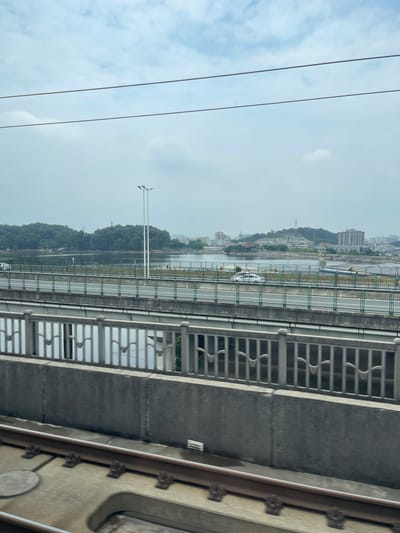 The image shows a view from a bridge or elevated structure, looking out over a river or body of water. On the opposite bank, a city or town is visible, with buildings nestled on a hillside covered in trees. A white car is driving on the bridge. The weather appears overcast or hazy. Several power lines or cables run across the top left of the frame, and the right side of the image shows part of the structure from which the photo was taken, including a metal railing and concrete. The orientation of the image is landscape, with the scene viewed from a slightly elevated perspective. The overall impression is one of urban development meeting natural scenery.