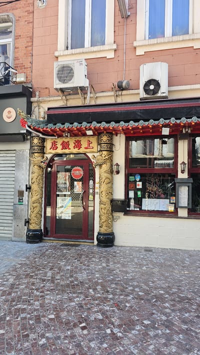 The image shows the entrance to a Chinese restaurant located in the city of Charleroi, Belgium. The restaurant has a distinctive Asian architectural style with a red and green tiled roof, ornate golden dragon-shaped pillars flanking the entrance, and a golden sign with Chinese characters above the door. The entrance itself is a glass-paned door with a dark red frame. Various stickers and notices are visible on the door, including "FRAGILE" and information about opening hours.
To the left of the entrance, there is a closed metal shutter, and a portion of a dark-colored building with a circular logo. Above the restaurant's entrance, there are two air conditioning units mounted on the facade of a brick building. One unit is a white Mitsubishi Electric unit, and the other is a white, older-style unit.
To the right of the entrance, there is a large window with a dark red frame, revealing the interior of the restaurant, which appears to be decorated in an Asian theme. Various menus and notices are displayed in the window. Next to the window, there is a black mailbox.
The ground in front of the restaurant is paved with small, reddish-brown and greyish-white square cobblestones arranged in a curved pattern. The lighting suggests it is daytime, with sunlight casting shadows. The overall scene depicts a commercial establishment in an urban environment.