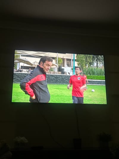 A television screen displays a scene with two men on a green lawn. The man closer to the viewer wears a black jacket with red accents and appears to be in discomfort or reacting strongly. The other man, further away and in a red shirt, seems to be throwing a ball. Buildings and trees form the backdrop, suggesting an outdoor or recreational setting.