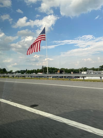 An American flag waves from a tall flagpole against a blue sky with scattered clouds. The flag is positioned in the upper left quadrant of the image. In the middle ground, there is a row of buildings and a parking lot filled with cars. A metal guardrail separates the parking lot from a highway in the foreground. The highway pavement is visible, with a white line marking the lane. The image appears to be taken from a moving vehicle, as evidenced by the motion blur on the road. The scene is set in Thurmont, United States of America.