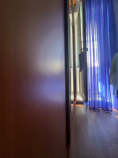 This image shows an interior view of a room with a wooden floor and blue curtains. On the left side of the image is a brown wall with a subtle purple reflection. To the right, a partially open window reveals blinds and blue sheer curtains. Sunlight streams through the window, casting shadows and highlights on the floor and curtains. A dark object, possibly a bag or garment, is hanging from the window frame. A light-colored garment is visible on the right side of the frame, hanging behind the curtains. The scene appears to be a residential interior, possibly a bedroom or living area, located in Muravlenko, Russia, given the provided context. There are no people visible in the image.