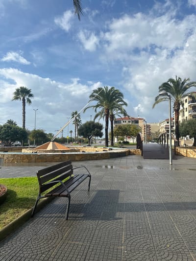 The image displays an urban public space in Pineda de Mar, Spain, under a partially cloudy blue sky during daytime. In the foreground, a dark-colored, slatted park bench with metal frame rests on the left side of a wet, gray paved pathway. Adjacent to the bench on the left is a small patch of green grass. The pathway features interlocking pavers with visible circular and square patterns, and appears wet from recent precipitation.

In the midground, a large circular stone fountain with visible water occupies the central-left area. At its center stands a light-colored, pyramidal structure from which a long, slender gnomon extends diagonally upwards, functioning as a sundial. Several tall palm trees are scattered throughout the scene, interspersed with smaller green trees and shrubs. To the right of the fountain, a dark, arched wooden pedestrian bridge with railings connects two sections of the paved area.

In the background, beyond a visible road with streetlights, multiple multi-story buildings with balconies are present, indicating a residential or commercial area. No individuals are clearly visible in the immediate foreground or midground. The overall scene suggests a coastal promenade or park area.