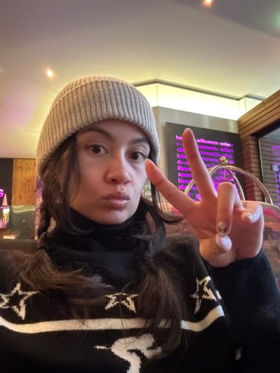 A selfie captures a young woman wearing a grey beanie and a black sweater with white star patterns. She makes a peace sign with one hand, holding it up near her face. Her expression is playful, with pursed lips. Behind her, a wall panel displays the word "welcome" in various languages, illuminated with a purple glow. A wooden structure and soft lighting are visible in the background.