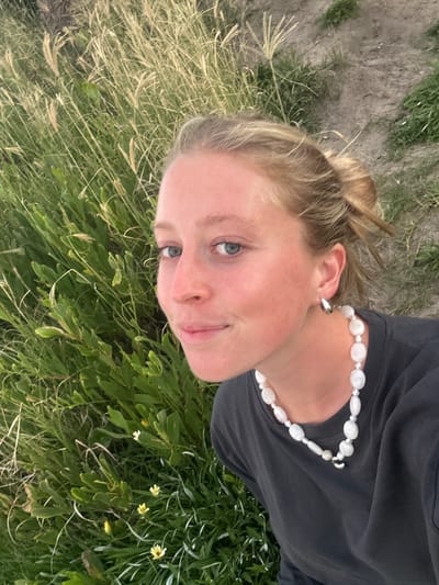 A young woman with fair skin and blonde hair in a bun is lying on her side in a field of green grass and vegetation with scattered small yellow flowers. She is wearing a black t-shirt and a pearl necklace. Her face is directed towards the camera. The background includes patches of brown soil. The image was likely taken outdoors during daylight hours. The specific location within Wollongong City Council, Australia, cannot be determined from the given image.