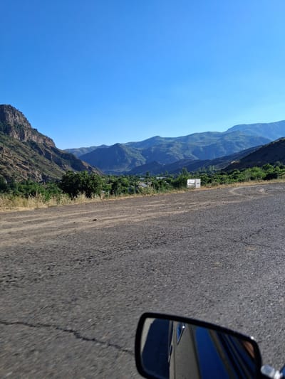 This media file captures a scenic view of a mountainous landscape with a clear blue sky. The perspective suggests the image was taken from a vehicle, as a car's side mirror is visible in the lower right corner. The road itself appears to be asphalt with some visible cracks and patches.
To the left of the road, a steep, rugged mountain range dominates the scene, covered in patches of green vegetation. The mountains stretch into the distance, creating a sense of depth and vastness. Below the mountains, a small settlement or village is visible, nestled amongst trees and greenery. A white sign with some text is also present near the roadside, though the text is not clearly legible.
The lighting and the bright blue sky suggest it is daytime, likely morning or afternoon. The weather appears to be clear and sunny, ideal for driving through this mountainous region. The location cues, such as the style of the mountains and the vegetation, are consistent with a setting in Shatin, Armenia. There are no people or specific activities depicted, emphasizing the natural beauty of the landscape. The overall mood is one of tranquility and exploration.