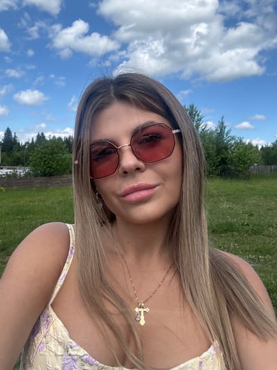A young woman with long, blonde hair and pink lipstick smiles as she takes a selfie outdoors. She wears pink-tinted sunglasses and a floral-patterned dress. A delicate gold necklace with a cross pendant rests on her collarbone. The background shows a lush green field, a treeline, and a bright blue sky with scattered white clouds, suggesting a sunny day. A rustic wooden fence is visible in the distance. The overall mood is relaxed and cheerful, indicative of a pleasant outdoor leisure activity. The image appears to have been taken during daylight hours, possibly in the afternoon, given the bright sunlight and clear sky. The location context is Пастухово, Russia. There is no visible text in the image.