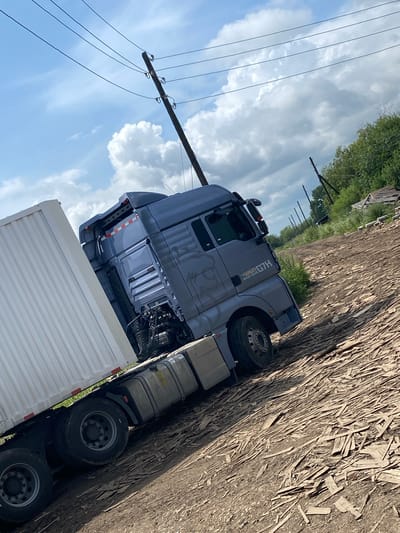 In Liubikha, Russia, on a dirt road covered with wood debris, a blue semi-trailer truck is parked. The truck is carrying a white trailer. Power lines stretch across the sky above. The scene appears to be in a rural or industrial area, with trees and dilapidated structures visible in the background. The sky is partly cloudy, with patches of blue visible.