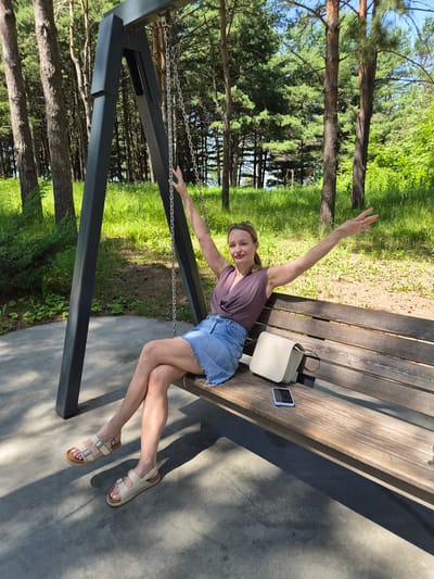 A woman with blonde hair, wearing sunglasses on her head, a mauve-colored V-neck top, and a denim skirt, is sitting on a wooden bench. She has her arms raised with her palms facing outward. Her legs are crossed at the knees, and she is wearing beige sandals. Next to her on the bench is a light-colored handbag and a smartphone. The bench is situated in front of a dark metal swing set. The background consists of a grassy area with trees, suggesting an outdoor park or recreational area. The scene is illuminated by bright sunlight, casting shadows on the ground. The location is identified as Arseniev, Russia.