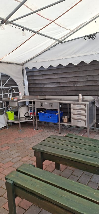 The image depicts an outdoor food preparation area under a white tent. The scene is located in Valma, Estonia. A stainless steel counter with a sink, faucet, and a built-in cooking surface is the central feature. Beneath the counter, a green plastic bucket, plumbing, and electrical outlets are visible. To the right, the counter has four drawers. A blue plastic crate sits on a shelf below the cooking surface. In the foreground, two dark green wooden benches are positioned on a paved brick patio. The background features a dark wooden fence. The tent's white fabric roof is supported by metal poles, and two Edison-style light bulbs hang from the structure. The overall impression is of a functional, open-air service area.