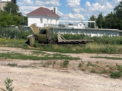 An abandoned olive-green truck sits in an overgrown lot under a partly cloudy sky. The truck's hood is propped open, revealing a rusted engine. The flatbed of the truck is also visible, with some wooden planks still attached. The truck appears to be in a state of disrepair and is surrounded by tall grass and weeds.
In the background, there is a white building with a brown roof and a tall fence. Beyond the fence, there are trees and more buildings, suggesting an industrial or rural area. The overall scene depicts a forgotten or neglected space, with the truck being the primary focus. The time of day is likely daytime, given the bright sunlight and shadows. The weather appears to be warm and possibly humid, with the fluffy clouds indicating fair weather.
There is no visible text in the image. The location context provided is "Челгаши, Kazakhstan."