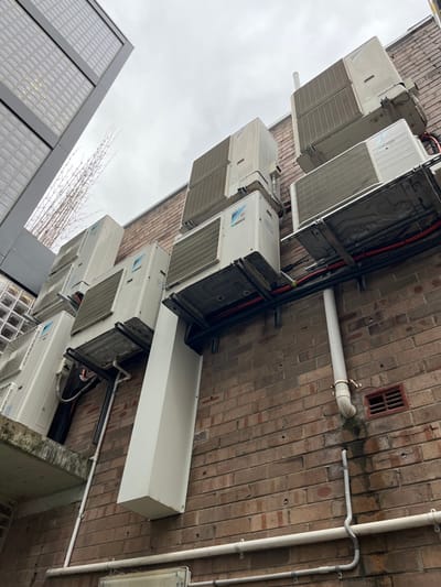 A row of air conditioning units are mounted on a brick wall, with pipes and electrical conduits running along the wall. The units are mostly white, with vents and various mechanical components visible. Some of the units are supported by metal frames. A white rectangular box is also attached to the wall beneath one of the units. A nearby building with glass brick windows and a grey frame can also be seen. The sky is overcast.