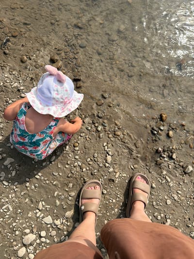 A child wearing a white sun hat with pink bows and a floral-patterned swimsuit sits on a pebble-covered riverbank in Rodnikovskaya, Russia. The child is playing with the pebbles near the shallow, clear water. The perspective is from above, showing the child's back and the legs of an adult wearing beige slide sandals and a brown skirt in the foreground. The sun is shining, creating reflections on the water's surface.