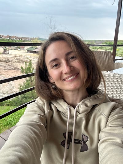 A woman is sitting in a wicker chair on a balcony overlooking a residential area of Zakharivka, Ukraine. She is wearing a light tan Under Armour hooded sweatshirt. The view from the balcony shows houses with brown rooftops and grassy areas and trees between buildings. The sky is overcast. The woman is looking at the camera and smiling. The balcony has dark metal railings.