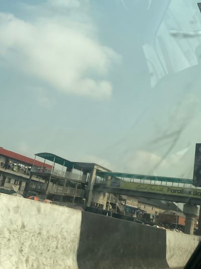 The image displays an urban scene in Road, Nigeria, under a light blue sky with scattered white clouds. In the foreground, a concrete barrier, painted white on the left and dark grey on the right, spans the lower portion of the frame. Above this barrier, a multi-story concrete building with a red corrugated metal roof is visible on the left, featuring laundry hung on an upper-level railing. To its right and extending across the mid-ground, a multi-level pedestrian bridge structure is prominent, constructed of concrete and metal with green corrugated canopies covering sections of its walkways. An advertising banner affixed to the bridge reads "Bank free with Parallex Ba" (likely "Parallex Bank"). Further urban buildings are partially visible beneath and behind the bridge. Reflections consistent with a view through a vehicle windshield are present in the upper right quadrant of the image. No distinct individuals or specific actions are identifiable.