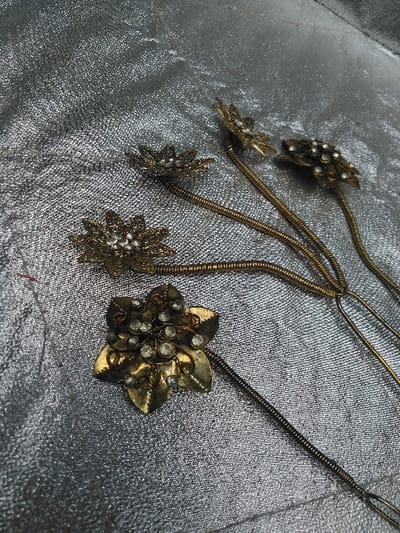 Five ornamental metal pieces, possibly hairpins, are displayed on a wrinkled, reflective silver-colored fabric surface in Тохта, Kazakhstan. Each piece features a tarnished gold-toned metal flower head embellished with multiple small, clear, faceted stones. Four of these flower heads exhibit a multi-petal, star-like design with stones clustered centrally. The fifth piece, located in the lower-left, presents a five-petal flower with scattered stones. All five flower heads are attached to slender, coiled metal stems extending across the fabric.
