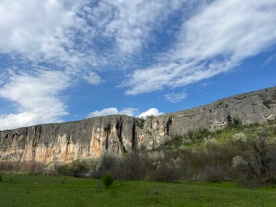 The image captures a broad, open landscape under a partly cloudy sky in Barkach, Bulgaria. A prominent feature is the large rock formation, a cliff or rocky slope, that stretches across the midground. The rock face has various colors: gray, beige, and brown. Below the rock, a line of trees and bushes are interspersed, marking a transition to a flat, grassy field in the foreground. The sky above has a mix of blue and white clouds, partially obscuring the blue sky. The general environment seems natural and rural. No human subjects are visible. The interactions between the natural elements of rock, trees, grass, and sky dominate the scene.