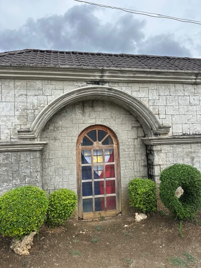 An outdoor, eye-level, medium shot captures a stone building facade in Oslob, Philippines, under an overcast sky. The building's exterior is constructed from light gray, rough-cut stones, arranged in a staggered pattern. A prominent arched architectural feature, also made of stone, is centered on the facade. Within this arch is a tall, narrow window with a wooden frame and divided panes. The window glass is tinted with the colors of the Philippine flag: red, blue, and yellow, creating a patriotic stained-glass effect. The roof above the facade is covered in dark, wavy tiles. In front of the building, on the ground, are three manicured, spherical green bushes. Two are smaller and positioned to the left of the window, while a larger one is to the right. The ground is covered in dirt, small stones, and sparse dry vegetation. Two power lines are visible in the upper portion of the image, stretching across the cloudy sky.