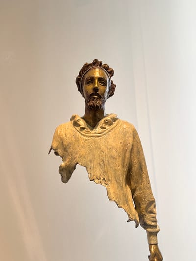This is a medium shot of a bronze sculpture of a man's bust, positioned against a plain, light-colored wall. The sculpture depicts the upper body of a man with detailed facial features, including a beard and curly hair. He is wearing a garment that appears to be a hooded robe or sweatshirt, which is unfinished and broken, giving the impression of being torn or dissolving. The texture of the bronze is visible, with a metallic sheen and variations in color from gold to brown. The lighting casts subtle shadows on the wall behind the sculpture, creating a sense of depth. The sculpture is set against a neutral background, emphasizing its form and texture. This sculpture is located in Honfleur, France.
