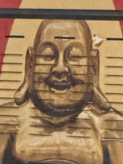 The image is a close-up, horizontally oriented, grainy photograph of a golden Buddha statue. The statue is depicted from the chest up, with its face prominently featured. Horizontal lines, resembling a measuring scale or a pattern, are superimposed across the image, passing over the statue. The background appears to be a textured yellow, with a sliver of red visible at the top left corner. There is a dark, thick horizontal band across the very top of the image. A small, irregular white object, possibly a piece of paper or a tag, is attached to the top right side of the statue's head. The statue's features, including its closed eyes, rounded cheeks, and serene expression, are visible. Its hands are not fully visible in the frame. The overall lighting is subdued, giving the statue a warm, aged appearance. There is no indication of the city of Shkotovo, Russia, within the image itself.
