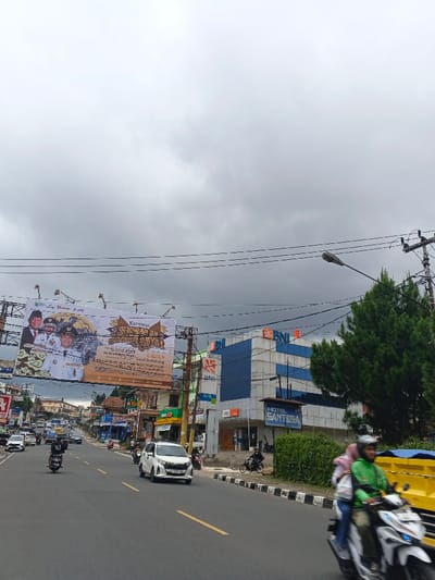 The image depicts a busy urban street scene in the city of Palayan, Philippines, under an overcast sky. The asphalt road, marked with yellow lines, accommodates a flow of vehicular traffic, predominantly motorcycles and cars.
On the right side of the street, a modern multi-story building with blue glass panels and white sections prominently displays multiple "BNI" logos. Below this, a sign for "HOTEL SANTOSA" is visible. This side of the road is bordered by a sidewalk with black and white striped curbs, lush green hedges, and tall, dark green pine-like trees partially obscuring the "BNI" building. Further down the sidewalk, a person is seen operating a small bicycle or scooter. In the immediate foreground, a motorcycle rider wearing a green helmet and jacket, accompanied by a passenger in a pink head covering, travels towards the camera. A partial view of a yellow truck is behind them.
On the left side of the street, a large billboard is suspended overhead, featuring images of several men in suits and traditional attire, along with text that includes "Karnaval Sumbar". A white sedan is visible in the middle lane, moving away from the camera, and another motorcycle with a single rider in dark attire is also moving in the same direction on the left lane. Further down the street, a continuous line of commercial buildings, some with awnings, recedes into the distance. Numerous utility poles with a dense network of overhead power lines crisscross the upper portion of the frame, and a street lamp extends from a pole on the right.