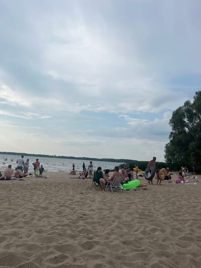 This image shows a crowded beach scene in Belarus, with many people enjoying a day by the water. The sand is a light brown, with ripples from the wind. The water is a murky blue, and there are trees in the background. The sky is overcast with some patches of blue.
The people on the beach are of various ages and are dressed in swimwear. Some are sitting on the sand, while others are standing or walking. There are several inflatable objects, including a large green ring and a tire. A few people are playing in the water.
The image appears to have been taken during the day, as there is natural light. The weather is warm and pleasant. The scene evokes a sense of leisure and enjoyment. There is no visible text in the image.