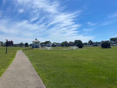 A wide, paved pathway curves through a lush green lawn, leading the viewer's eye towards a picturesque scene under a bright blue sky dotted with wispy white clouds. In the distance, a white gazebo with an ornate roof stands prominently, surrounded by a collection of charming houses with varied architectural styles. Two circular fountains with water jets are visible on the grass, adding a refreshing element to the park-like setting. Cars are parked along the edge of the green, suggesting a public space, perhaps a park or a town common. The architecture of the houses, with their gabled roofs and varied colors, hints at a coastal or historic New England town. The bright sunlight and clear sky indicate a pleasant day, likely daytime. There are no visible people engaged in any specific activity, contributing to a sense of peacefulness and tranquility. The overall impression is one of a well-maintained public park in a quaint American town, possibly during the summer months due to the vibrant green of the grass.