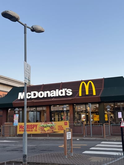 The image captures a McDonald's restaurant exterior under a clear, pale blue sky. A tall grey lamppost with two weathered, oval-shaped lights stands prominently in the foreground to the left. A parking sign, reading "2 Hours Free Parking," is attached to the lamppost.

The McDonald's building features its iconic golden arches and white "McDonald's" signage. A large banner advertising a "McCRISPY" burger with cheese and bacon is displayed on the left side of the storefront. The restaurant's facade consists of large windows and red brickwork.

In front of the restaurant, there is a paved area with a pedestrian crossing. A small white disposable cup is visible on the ground near the crossing. The overall environment appears to be an urban setting, likely a commercial area, given the presence of the large fast-food chain and the parking facilities. The lighting suggests it is either early morning or late afternoon, with the sun casting long shadows. The location is identified as Boston, United Kingdom, based on the context provided.