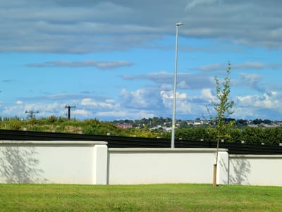 The image depicts an outdoor scene on a sunny day with scattered clouds. In the foreground, a neatly manicured green lawn stretches out. A white, textured wall with dark green horizontal panels along the top forms a barrier. A young tree, supported by a wooden stake, stands to the right of the wall. Behind the wall, a gentle slope covered in lush green vegetation leads up to a distant town or village. Silhouetted against the sky are power lines and telephone poles on the left, and a tall street lamp stands to the right of the young tree. The town in the distance features buildings with red roofs and other structures, nestled amongst trees. The sky is a vibrant blue, dotted with fluffy white clouds interspersed with darker, elongated grey clouds. The overall impression is one of a well-maintained, possibly residential or developing area, with a pleasant, slightly breezy atmosphere suggested by the dynamic cloud formations. No people are visible in the image, and there is no visible text. The lighting and cloud cover suggest it is daytime, likely late morning or early afternoon. The location context provided indicates Waterford, Ireland.