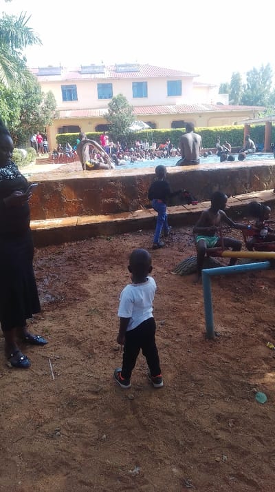 This image depicts an outdoor recreational area in Siaya Township ward, Kenya. In the immediate foreground, a young child wearing a white t-shirt, black trousers, and sneakers stands on reddish-brown dirt ground, facing away from the camera towards a swimming pool. To the child's left, a woman in a black dress or skirt and top holds a smartphone, partially obscured by the left frame. Further right, another child in blue jeans and a dark top walks rightward, holding a red item. A third child, wearing green swim trunks, sits on a colorful seesaw-like playground structure adjacent to a black tire.
Behind a low, textured concrete wall, a crowded swimming pool is visible, filled with numerous individuals, primarily children, engaged in swimming and water activities. Several adults are also present around the pool's edge and in the water. An adult male sits on the pool's edge with his back to the camera.
In the background, a multi-story building with a light-colored facade and a reddish-brown tiled roof is prominent. Three solar water heaters are installed on the roof, and several blue-framed windows are visible on the upper floors. Green trees and bushes surround the building and the pool area, indicating a vegetated environment under a bright, possibly overcast, sky.