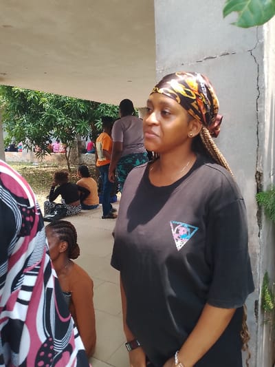 The image depicts a group of people in an outdoor setting, possibly within the city of Urualla, Nigeria. A woman with braided hair, wearing a black t-shirt and a patterned headscarf, stands prominently in the foreground, looking towards the right. In the background, other individuals are seated or standing, some appearing to be waiting. The location seems to be a covered area, possibly a building or a structure with a roof, adjacent to some trees and vegetation. A concrete pillar with cracks is visible on the right. The scene suggests a casual gathering or waiting area.