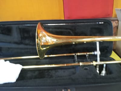 The image shows a brass trombone lying in its open black case. The trombone is golden and reflects the light. The inside of the case is black and appears to be velvet or a similar soft material, shaped to hold the instrument. A white cloth or fabric is visible on the left side, possibly used for cleaning or protecting the instrument. The case is placed on a surface that is partly visible, revealing a yellow and red background, suggesting it might be a room or a studio. There are no people visible in the image. The lighting appears to be indoor lighting, creating reflections on the polished brass. There is no visible text. The scene conveys a sense of readiness, as if the instrument is about to be played or has just been put away. The specific location cues are limited to the colors in the background.