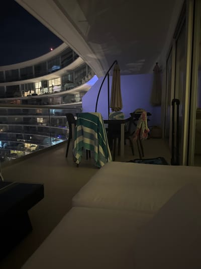 A balcony scene overlooking a cruise ship at night. A striped towel hangs over a chair near a closed umbrella. Other furniture includes a table, stacked chairs, and a white cushioned lounge chair. Across the ship's exterior, rows of lighted cabins are visible against the dark sky.