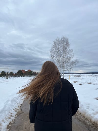 The scene, located in Kalinino, Russia, captures an individual walking away from the camera on a narrow, dark-colored path. The person has long, light-brown hair, visibly blowing to the left, and is wearing a black puffer jacket with horizontal stitching. The path is bordered by snowbanks on both sides. To the right, an expansive, snow-covered field with patches of exposed dry vegetation stretches towards a distant horizon marked by a faint tree line. A single bare deciduous tree with light-colored bark stands prominently in the mid-ground within the field. In the left background, several residential buildings, including one with a reddish-brown roof, are visible behind a fence and sparse trees. The sky is entirely overcast with a uniform layer of gray clouds. The environment indicates a cold, winter or early spring season.