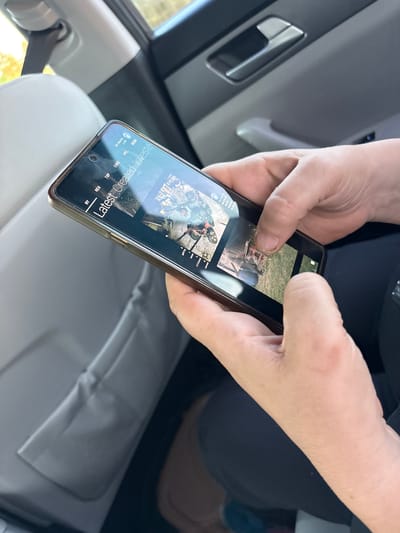 A close-up shot captures a person's hands holding and operating a smartphone inside a vehicle. The phone's screen displays a grid of images, with the text "Latest Created Investe" visible. The background suggests the interior of a car, with a grey headrest, seatbelt, and door handle. The lighting indicates it is daytime, and the scene appears to be in a residential area. There are no discernible landmarks or specific details pointing to the city of Slaveevo, Bulgaria.