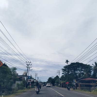 The image presents a daytime view down a two-lane asphalt road in Kabat, Indonesia, under an overcast sky. The road features a yellow centerline and is flanked by dense tropical vegetation, including large trees and tall palm trees. Numerous overhead power lines are strung across utility poles, creating a web-like pattern against the sky.
On the left side of the road, beyond the trees, the distinctive domes and spires of a mosque are visible. Adjacent to the mosque, various buildings, likely houses or small shops, line the roadside. A yellow triangular warning sign is partially visible in the foreground on the left, and a blue pedestrian crossing sign depicting a walking figure is also seen near a utility pole.
The right side of the road features more lush trees and several low-rise structures, possibly roadside businesses, one with a red-tiled roof and bamboo scaffolding. A vertical banner is affixed to one of these structures, with the word "REZHA" prominently visible, alongside other text in Indonesian.
Traffic is active on the road, with multiple motorcycles and a white car heading away from the viewer. A person on a scooter is in the immediate foreground, and other motorcyclists are further down the road. The scene captures a typical, bustling roadside environment in a Southeast Asian setting, characterized by the interplay of natural elements, local infrastructure, and daily activity.