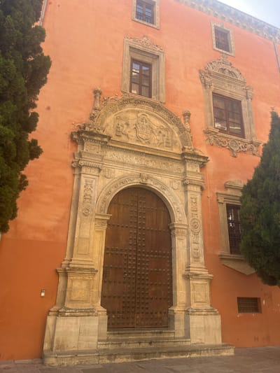 The image shows the ornate facade of a building in Granada, Spain, specifically the main entrance of the Corral del Carbón. The building's exterior is painted a vibrant orange hue. The central focus is a large, arched wooden doorway, intricately decorated with metal studs. Flanking the doorway are elaborately carved stone columns and pediments, featuring decorative elements and what appear to be heraldic symbols above the arch. Several windows with dark frames are visible on the upper levels of the building, some with decorative stone surrounds. Two large, dark green evergreen trees frame the entrance, partially obscuring the building on the left and right sides. The ground in front of the entrance is paved with stone tiles. The lighting suggests it is daytime, with shadows cast by the architectural elements and the trees.