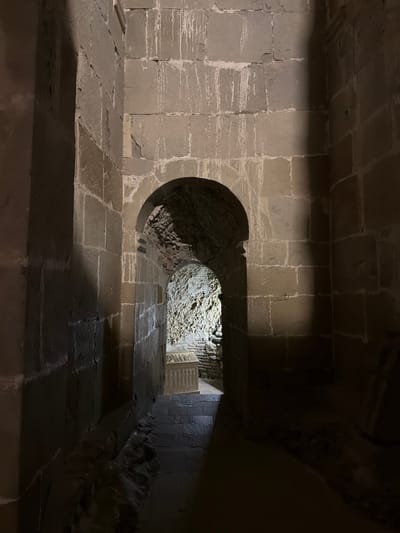 The image depicts an interior view of a stone structure, possibly an ancient building or tomb, with an arched passageway leading to another area. The walls are constructed of large, irregular stone blocks, with visible mortar between them. The stonework is weathered and shows signs of age, including some staining and erosion.

The foreground shows a dark, narrow corridor with a stone floor. To the right, the wall is illuminated by a light source, casting a shadow. Ahead, an arched opening reveals a dimly lit space beyond. This inner area features rough, uneven rock walls and what appears to be a stone staircase leading down. A sarcophagus-like object with carved details is visible in the background, illuminated by a light source. The overall atmosphere is ancient, solemn, and somewhat mysterious.

The specific location in the city of Unknown, Georgia, is not identifiable from the visual information presented in the image.