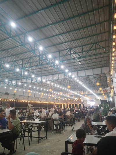 The image shows a crowded, spacious covered dining area, likely a restaurant or food hall, located in Namplohbaro, Indonesia. The structure features a high, gabled roof with corrugated metal sheeting supported by exposed teal-colored metal trusses. Overhead lighting consists of numerous bright white LED lights attached to the trusses, supplemented by decorative string lights with warm-toned bulbs and hanging wicker-style lanterns distributed throughout the space. The floor is a light-colored concrete.
Multiple square and rectangular tables with dark surfaces and black metal legs are arranged across the floor, each surrounded by black chairs. Approximately 50 to 70 individuals, predominantly adults, are seated at these tables. Many women are wearing hijabs. People are observed eating, drinking, and engaging in conversation in a casual setting.
In the mid-ground, a decorative wall segment features several illuminated arched alcoves. Above this, a horizontal display band shows framed pictures. Towards the right side of the image, a "KASIR" (Cashier) sign is visible, indicating a service point, next to some potted green foliage. The overall scene depicts a busy, well-lit, and active communal dining environment.
